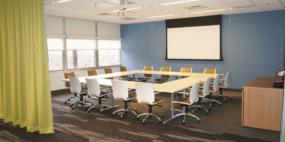 Conference room with a window, a screen, a curtain, and 16 chairs around a square table.